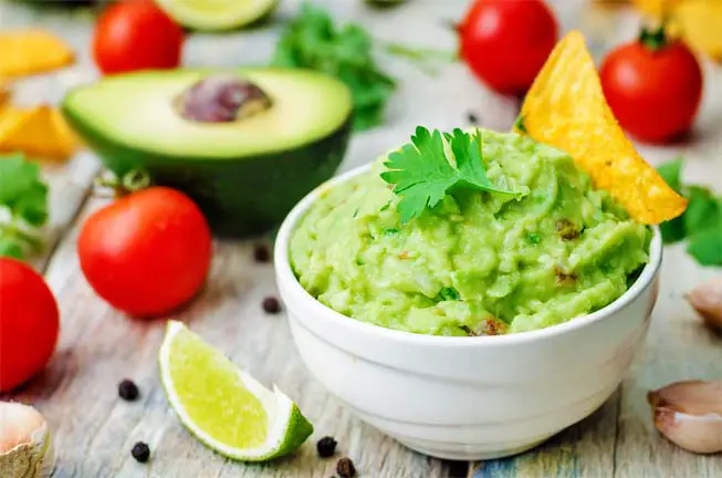 Heaping bowl of homemade guacamole by tomatoes, lime, garlic, avo slice, herbs.