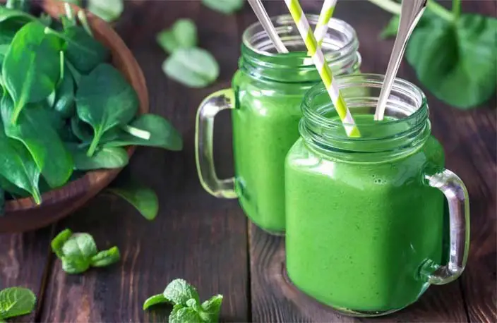 Two spinach smoothies on a rustic board by fresh spinach leaves.