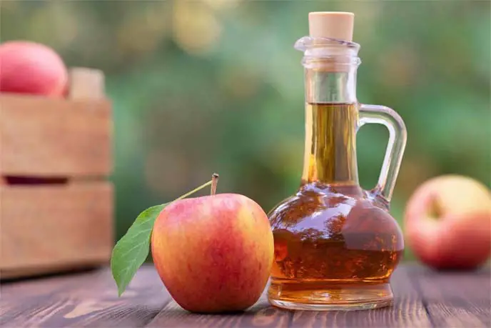 Jug of rich apple cider vinegar propped against a natural landscape on a wooden bench by fresh whole organic apples.