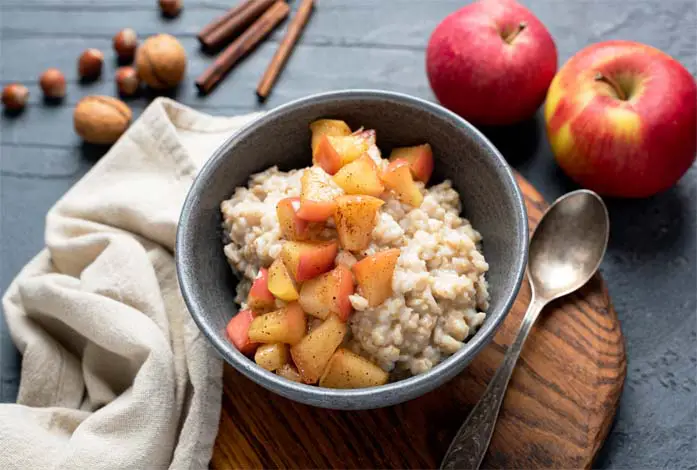 Top view of rich succulent porridge in a bowl by whole apples, cinnamon sticks, walnuts, hazelnuts, and an oversized cloth napkin positioned on a board.