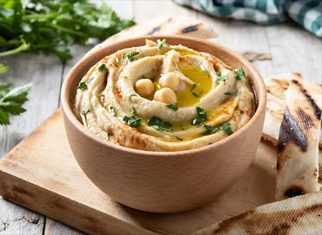 Bowl of delicious homemade hummus on a cutting board with pita bread and fresh herbs.