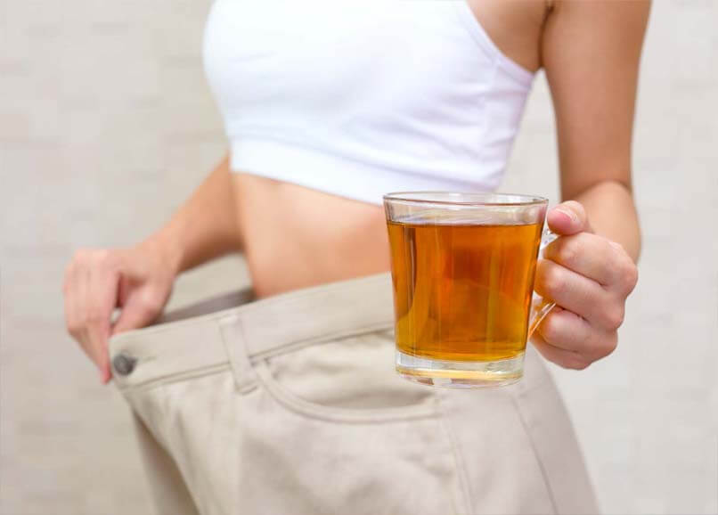 Girl holding a mug of tea displays weight loss by showing her shrunken waistline in oversize pants.    