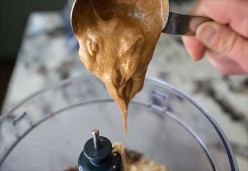 A person tilting a scoop of almond butter over a blender. 