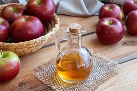 Glistening jug of rich apple cider vinegar on a wooden table by a basket of Red Delicious apples and assorted crisps.  