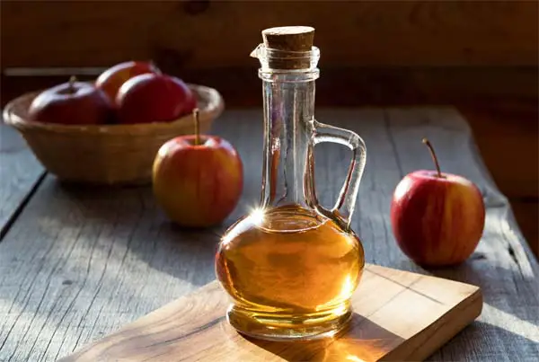 Glistening jug of golden apple cider vinegar on a rustic table by whole fresh  apples in shadowy sunlight.