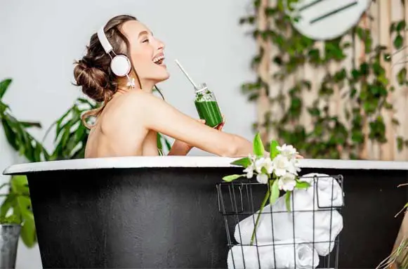 Happy smiling girl listening to music in the bathtub and drinking barley grass juice. 