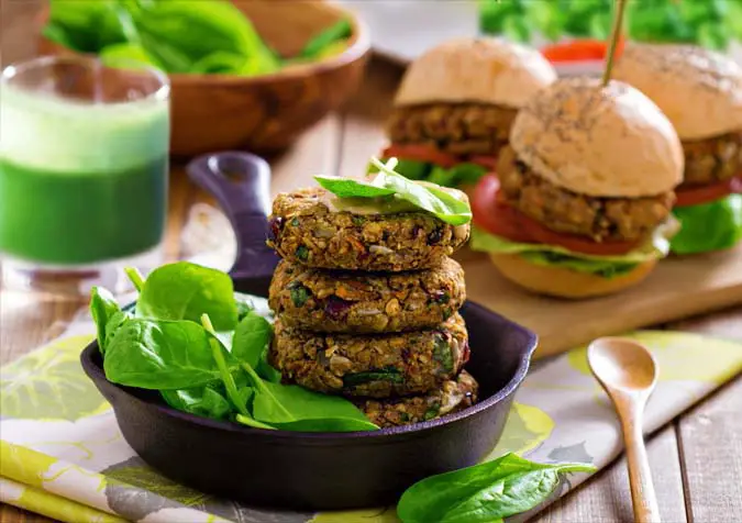 Delicious assortment of vegan black bean burgers with plump tomatoes, avocado, and greens by a stack of patties and spinach leaves in a cast iron pan. 