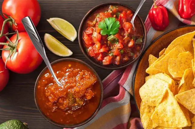 Top view of festive organic chips and bowls of chunky homemade salsa displayed by fresh whole tomatoes, peppers, avocados, and sliced lemon.