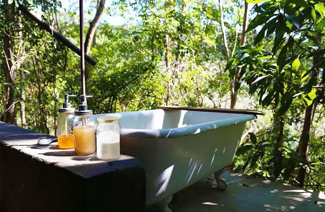 A large Clawfoot bathtub situated on an outdoor platform hugged by trees and giant jars of Castile and Epsom salts. 