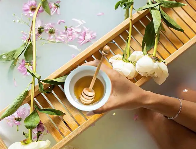 Top view of a girl's hand in a rose petal Epsom salt bath holding chamomile tea ointment above a caddy stacked with long-stemmed roses.