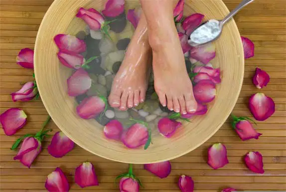 Top view of a woman's feet crossed in a soothing Epsom salt foot soak with rose petals and river rock in a bamboo basin.