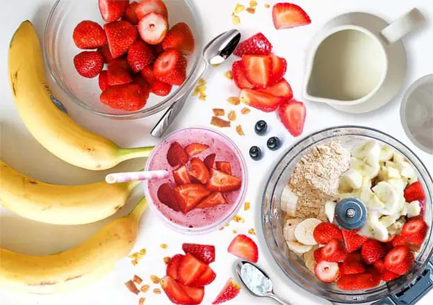 Strawberry banana smoothie recipe in progress with stacked blender, strawberries, bananas, ice, almond milk.