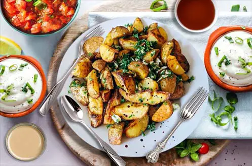 Top view of delicious spicy potato wedges on a cutting board plate by Ranch, tahini, salsa, and lava sauce bowls. 