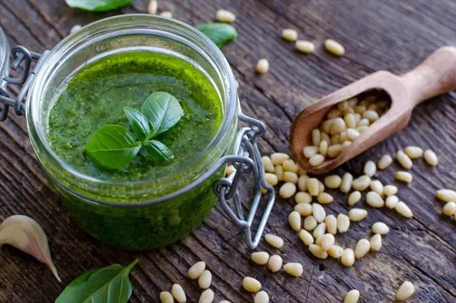 Open mason jar of succulent vegan pesto by a scoop of pine nuts scattered across a board. 