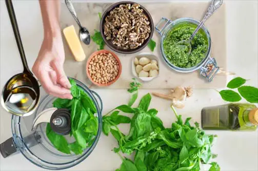 Overhead of a woman's hand prepping a delicious vegan pesto recipe with basil, pine nuts, garlic, olive oil, and herbs. 