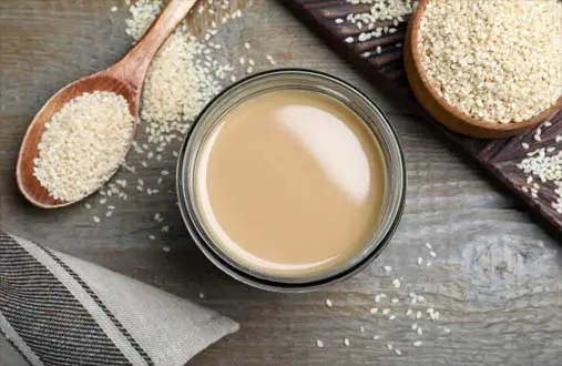 Top view of creamy smooth tahini sauce by an overflowing bowl and spoonful of raw organic sesame seeds. 