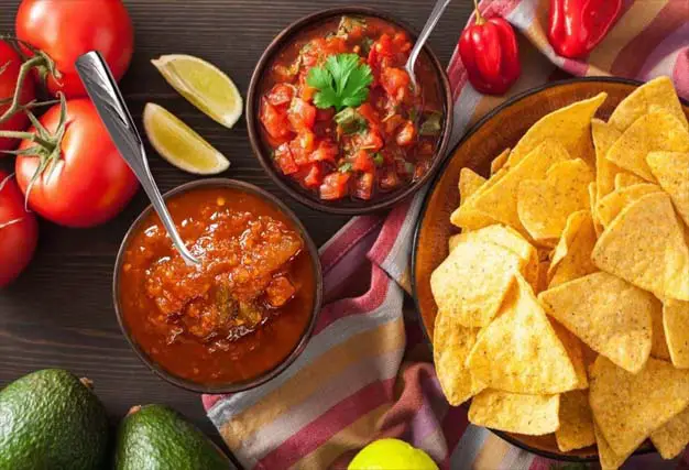 Top view of festive organic chips and bowls of chunky homemade salsa displayed by fresh whole tomatoes, peppers, avocados, and sliced lemon.