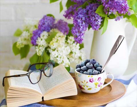 A cup and spoon of fresh blueberries by an open book topped with reading glasses and surrounded by vases of wildflowers.
 