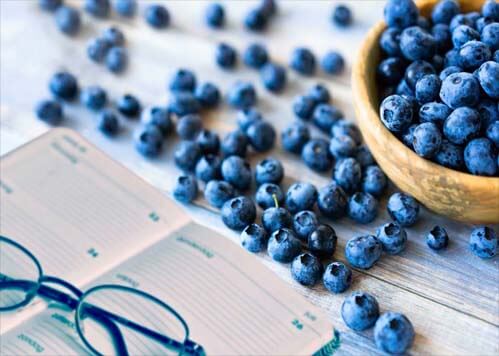 An overflowing bowl of blueberries by an open scheduling book and reading glasses.