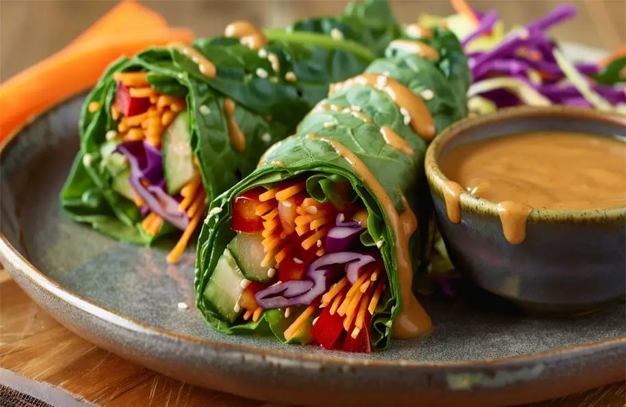 A plate of delectable vegan lettuce wraps by a bowl of creamy rich peanut sauce and organic veggies. 