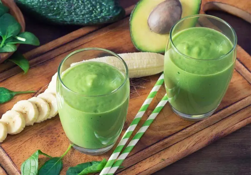 Top view of two avocado smoothies by bananas, spinach, and sliced avocado. 