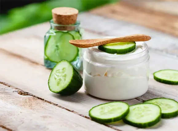 A Jar of cucumber facial cream and tiny wooden spoon by sliced cucumbers on a rustic board.  