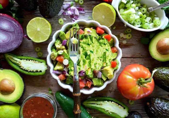 Top view of homemade guacamole  bowl with tomatoes, limes, onions, salsa, chili peppers, and avocados. 