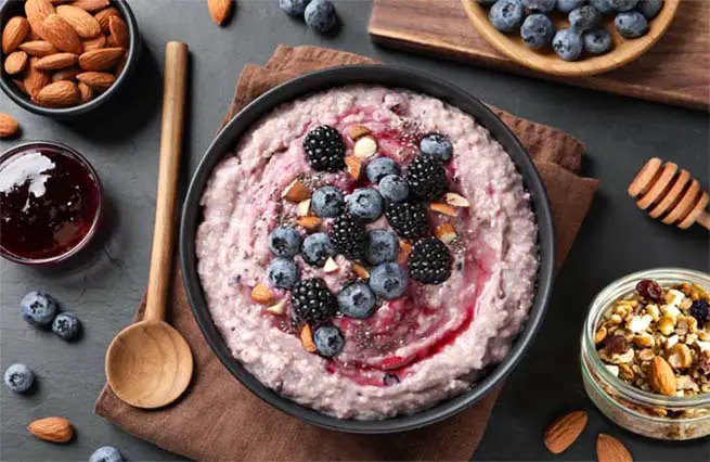 Top view of rich vegan Himalayan Tartary Buckwheat porridge in a bowl stacked with berries and nuts  surrounded by almonds, granola, blueberries, and maple syrup.