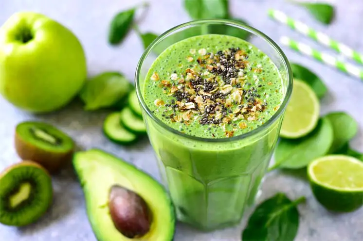 Fresh green smoothie topped with seeds and surrounded by avocado, limes, apples.  