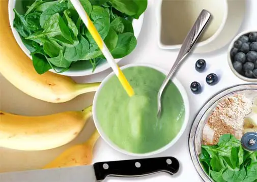 Top view of spinach smoothie ingredients with stacked blender, leaves, bananas, plant milk, blueberries. 