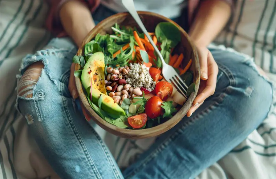 Top view of a woman in faded jeans holding a large wooden bowl of nutritious vegan foods.   