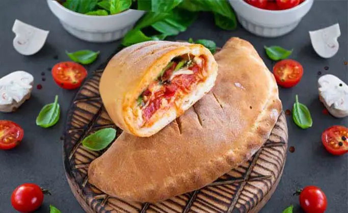 Rich vegan calzone with Himalayan Tartary Buckwheat crust surrounded by tomatoes, mushrooms, and spinach leaves on a bamboo board.