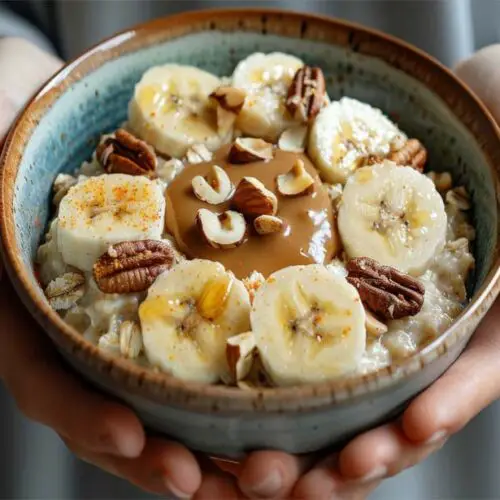 A man's hands cupping a stacked bowl of high-protein quinoa breakfast porridge topped with banana, almond butter, and mixed nuts.