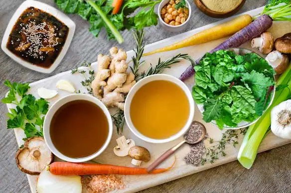 Top view of broth, veggies, herbs, and a wide array of vegan bone broth ingredients on a slanted cutting board.