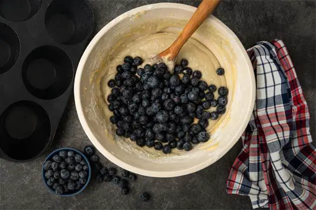 Top view of Himalayan Tartary Buckwheat muffin batter covered with fresh blueberries by a cast iron muffin pan, a bowl of more berries, a plaid kitchen towel, and an inserted wooden spoon ready to stir. 