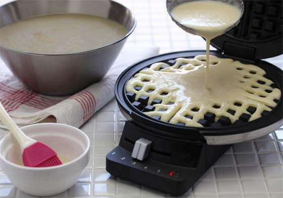 Close-up of a bowl of batter, a basting brush in a bowl, and a ladle pouring Himalayan Tartary Buckwheat waffle mix into a hot waffle iron on setting 3.  