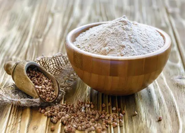 A wooden bowl stacked with Himalayan Tartary Buckwheat flour by a scoop and pile of Tartary seeds.