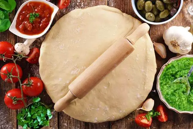 Top view of raw Himalayan Tartary Buckwheat pizza dough flattened on a board beneath a rolling pin and surrounded by tomatoes, pizza sauce, garlic, pesto, olives, and greens.   