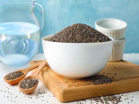 Stacked bowl and spoons of chia seeds on a wooden cutting board by small mixing containers and a pitcher of water. 