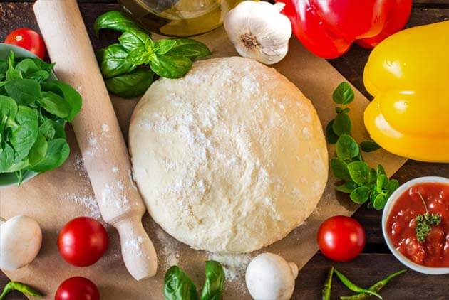 Top view of Himalayan Tartary Buckwheat pizza dough in a ball surrounded by tomatoes, garlic, mushrooms, bell peppers, greens, pizza sauce, and a rolling pin.