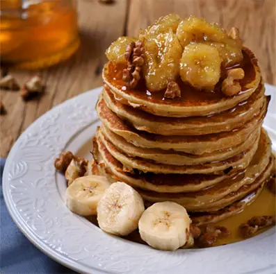 Tall stack of Himalayan Tartary Buckwheat pancakes with fresh and glazed bananas, walnuts, cashews, and maple syrup. 