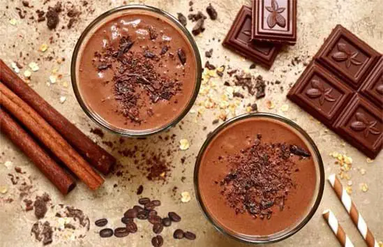 Top view of rich chocolate coffee protein smoothies topped with cacao and surrounded by chocolate bars, cinnamon sticks, and oats.  