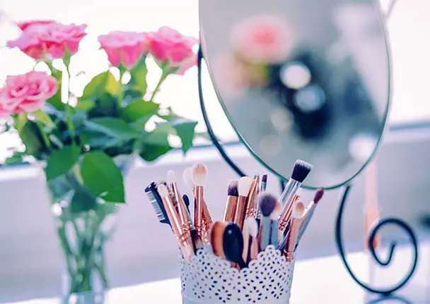 Vase of natural beauty products on a sunlit counter by fresh pink roses and a vanity mirror.