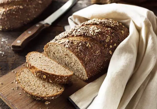 Sliced loaf of rich Himalayan Tartary Buckwheat bread on a cutting board by a knife and hugged by a kitchen towel warmer.