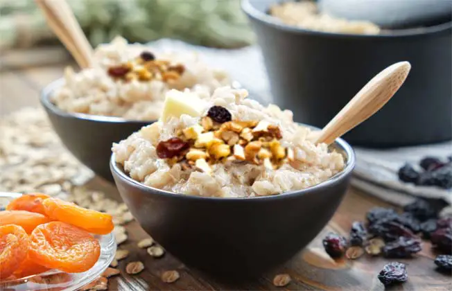 Two stacked bowls of Himalayan Tartary Buckwheat porridge topped with nuts and dried fruit beside a large cast iron pot and raisins, apricots, oats on a picnic table. 