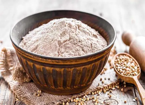 Large bowl of Himalayan Tartary Buckwheat flour by a spoon of seeds and rolling pin on burlap.