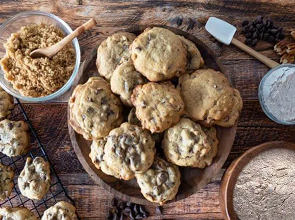 Plate of Himalayan Tartary Buckwheat chocolate chip cookies surrounded by flour, cookie batter, date sugar, cacao chips, nuts, and a spatula on a wooden board.