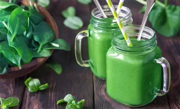 Two giant mugs of rich creamy green smoothie by a bowl of spinach on a rustic board.