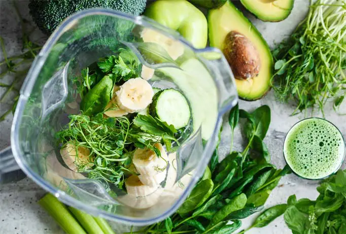 Top view close-up of blender stacked with greens, sprouts, and bananas surrounded by more greens, an avocado, celery, a wheatgrass shot.   