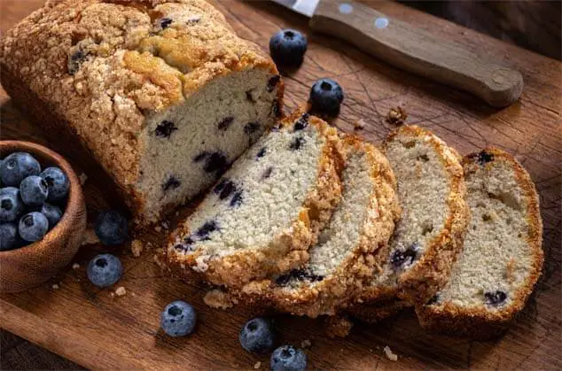 Sliced loaf of rich Himalayan Tartary Buckwheat blueberry bread by fresh blueberries on a cutting board.